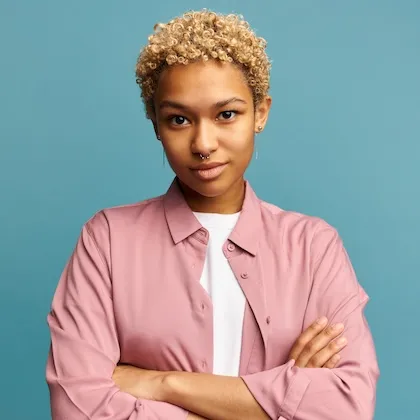 Confident young person with curly blonde hair, wearing a pink shirt over a white top, standing with arms crossed against a blue background.