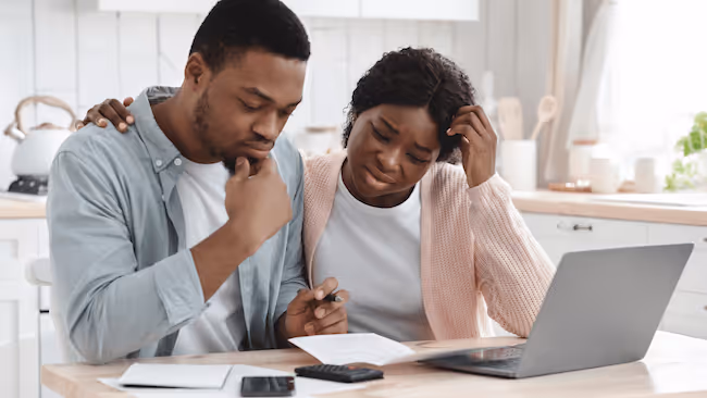 stresed couple in their kitchen, looking at a computer and paperwork