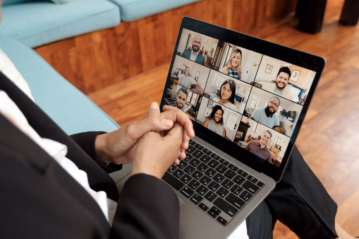 a well-dressed person sites on a couch with their laptop, as they conduct a video call with 9 other employees