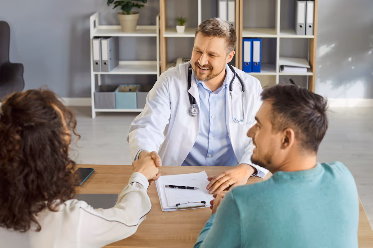 A physician shakes hands with a woman across a desk, as her male partner looks on.