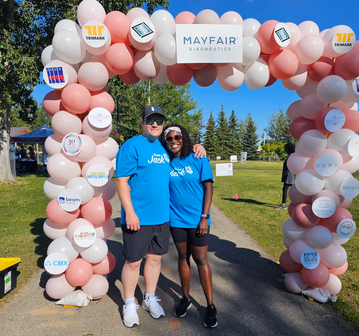 Kayann smiling and posting with someone on a nice day, under a balloon arch marked Mayfair Diagnostics