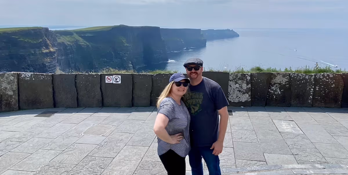 Vacation photo of Alicia and her husband, pictured on top of a cliff, probably somewhere in the North Atlantic, like Ireland or Newfoundland or Iceland or the Azores