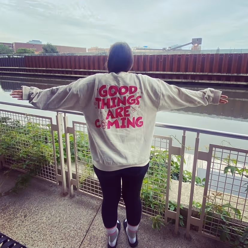 Jenn poses overlooking a river with her back to the camera. Her sweater says Good Things Are Coming.