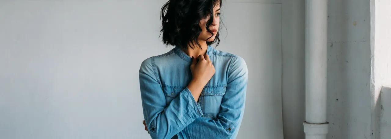 Woman with short dark wavy hair wearing a denim shirt, standing thoughtfully against a light gray wall