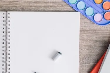 Overhead view of an open blank spiral notebook on a wooden desk surrounded by watercolor paints, an eraser, a phone, and a small plant
