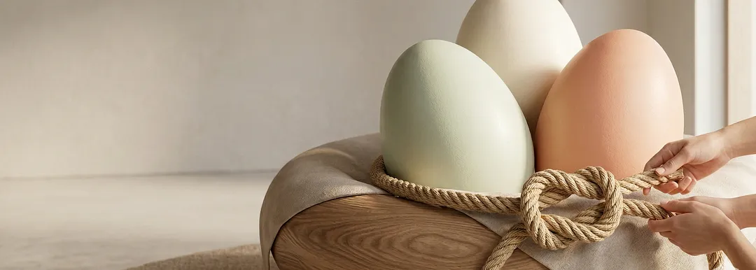 Three gigantic eggs in sage green, terracotta, and cream displayed on a wooden platform with rope, while hands tie a knot in the foreground