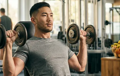 Man in a grey t-shirt doing dumbbell shoulder presses in a well-lit gym. There's fruit in the background. 