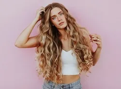 Woman with long wavy highlighted hair posing against a pink wall, wearing a white crop top and jeans