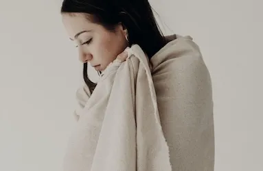 Woman with dark hair wrapped in a cream linen blanket, looking downward against a soft neutral background