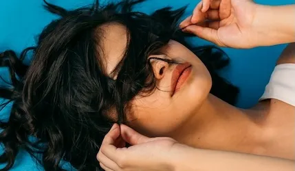 Woman with dark curly hair spread around her face lying on a blue background, hair partially covering her eyes