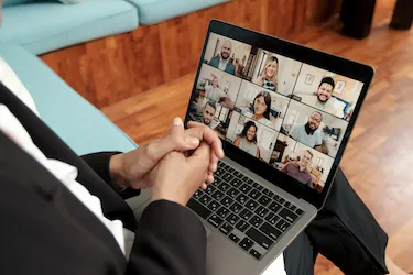 a well-dressed person sites on a couch with their laptop, as they conduct a video call with 9 other employees
