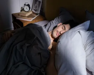a man appears to be sleeping peacefully. the light shines in from a window. a stereotypical analog alarm clock is on the bedside table.