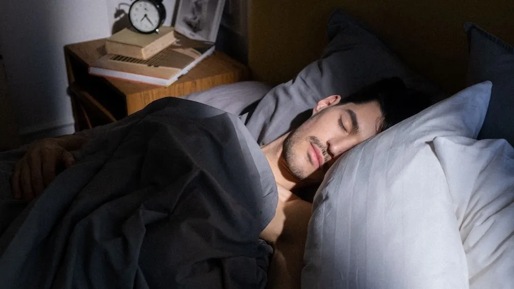 a man appears to be sleeping peacefully. the light shines in from a window. a stereotypical analog alarm clock is on the bedside table.