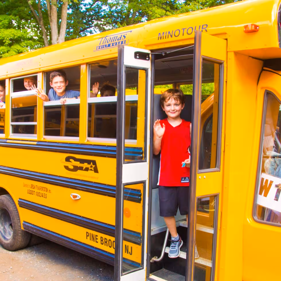 Children waving from the windows and entrance of a yellow school bus parked outdoors with green trees in the background.