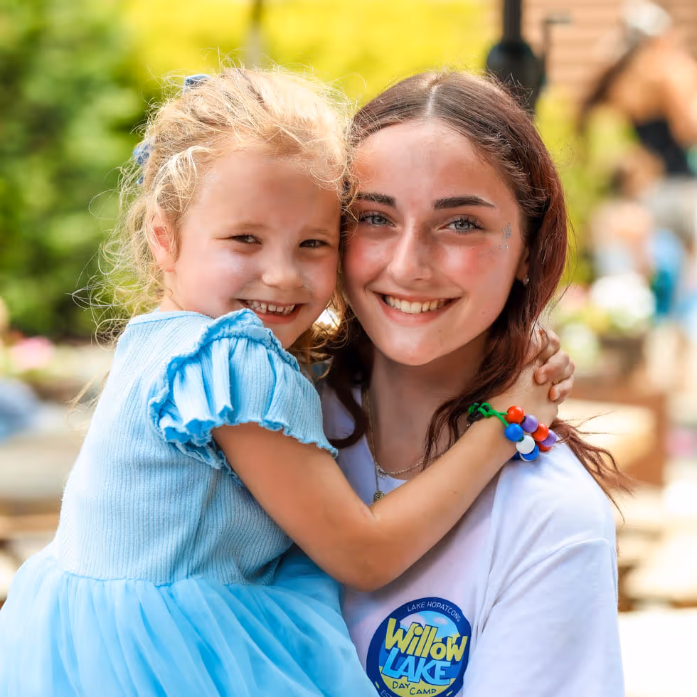 Smiling female counselor holding a young girl wearing a blue dress outdoors at Willow Lake Day Camp.