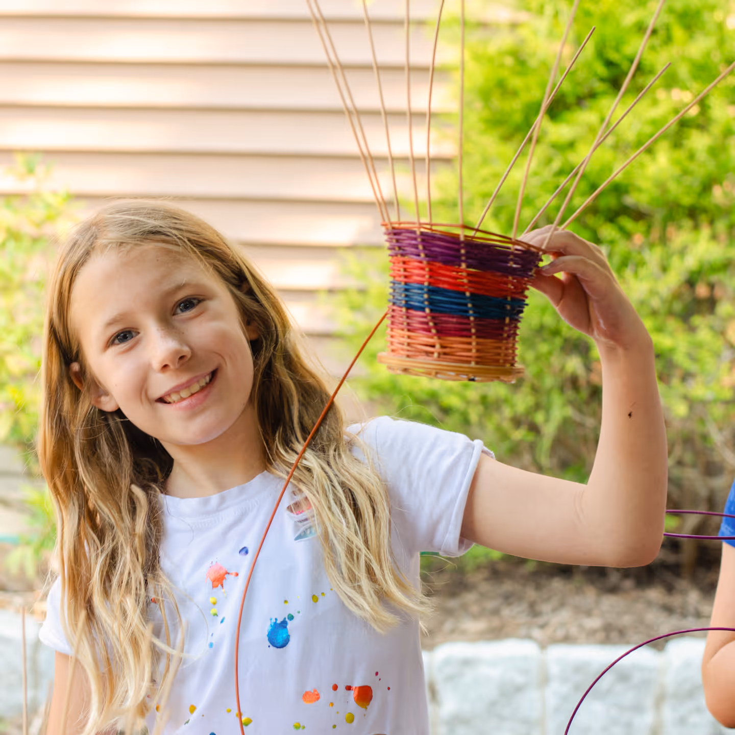 Smiling young girl holding a colorful woven basket in a garden setting.