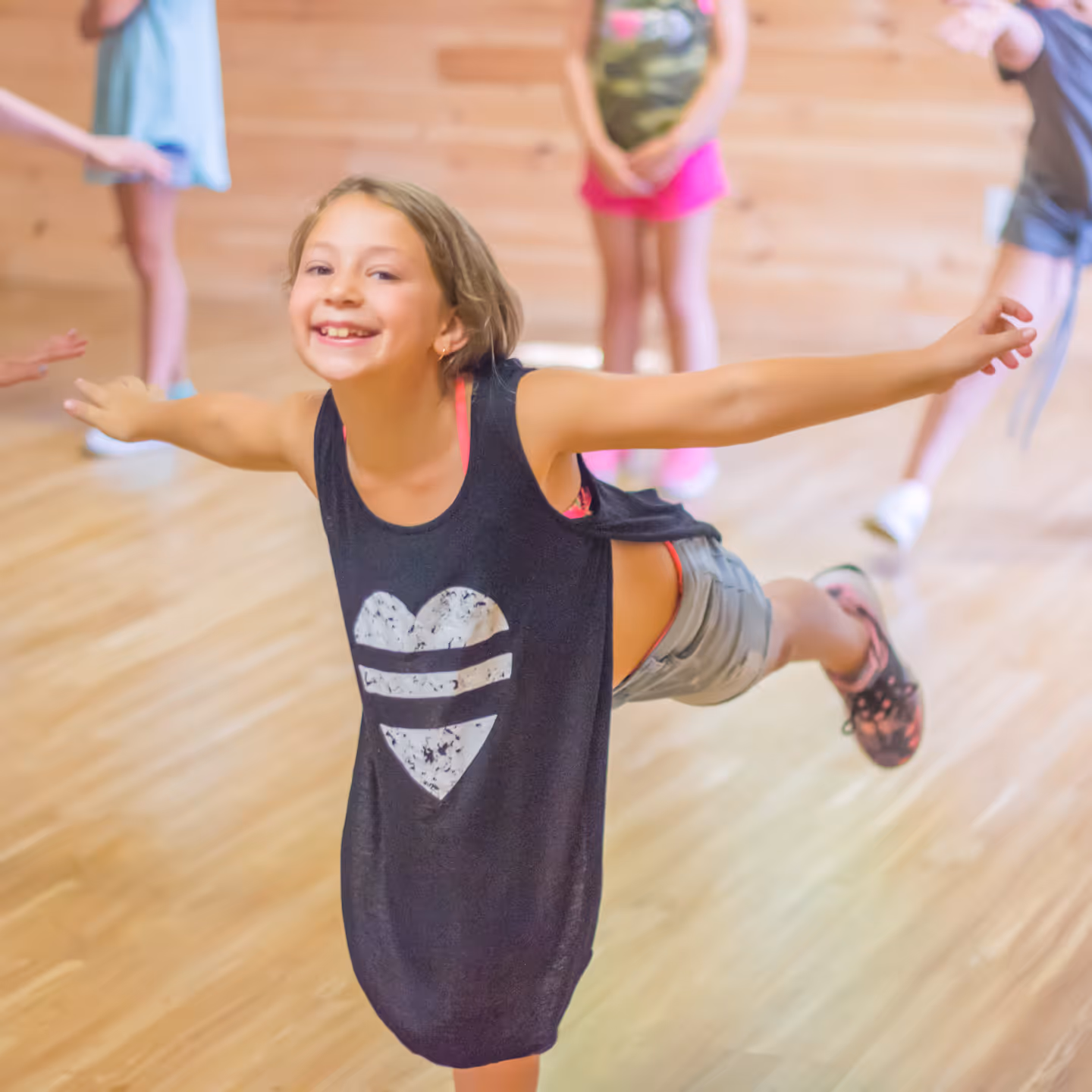 Smiling girl in a black tank top balancing on one leg with outstretched arms in a dance studio.