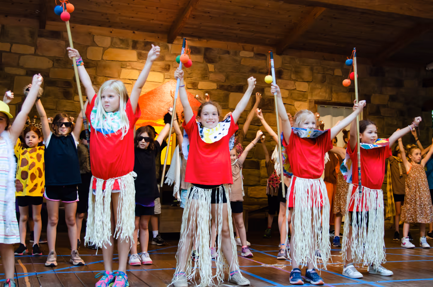 Group of children dressed in colorful costumes with arms raised, performing on stage with a stone wall background.