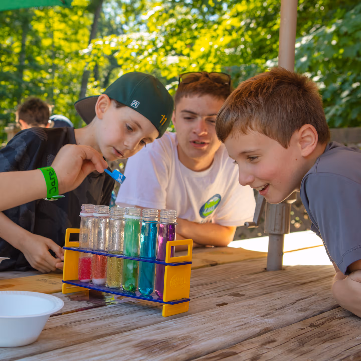 Three boys outdoors conducting a colorful science experiment with test tubes filled with liquids on a wooden table.