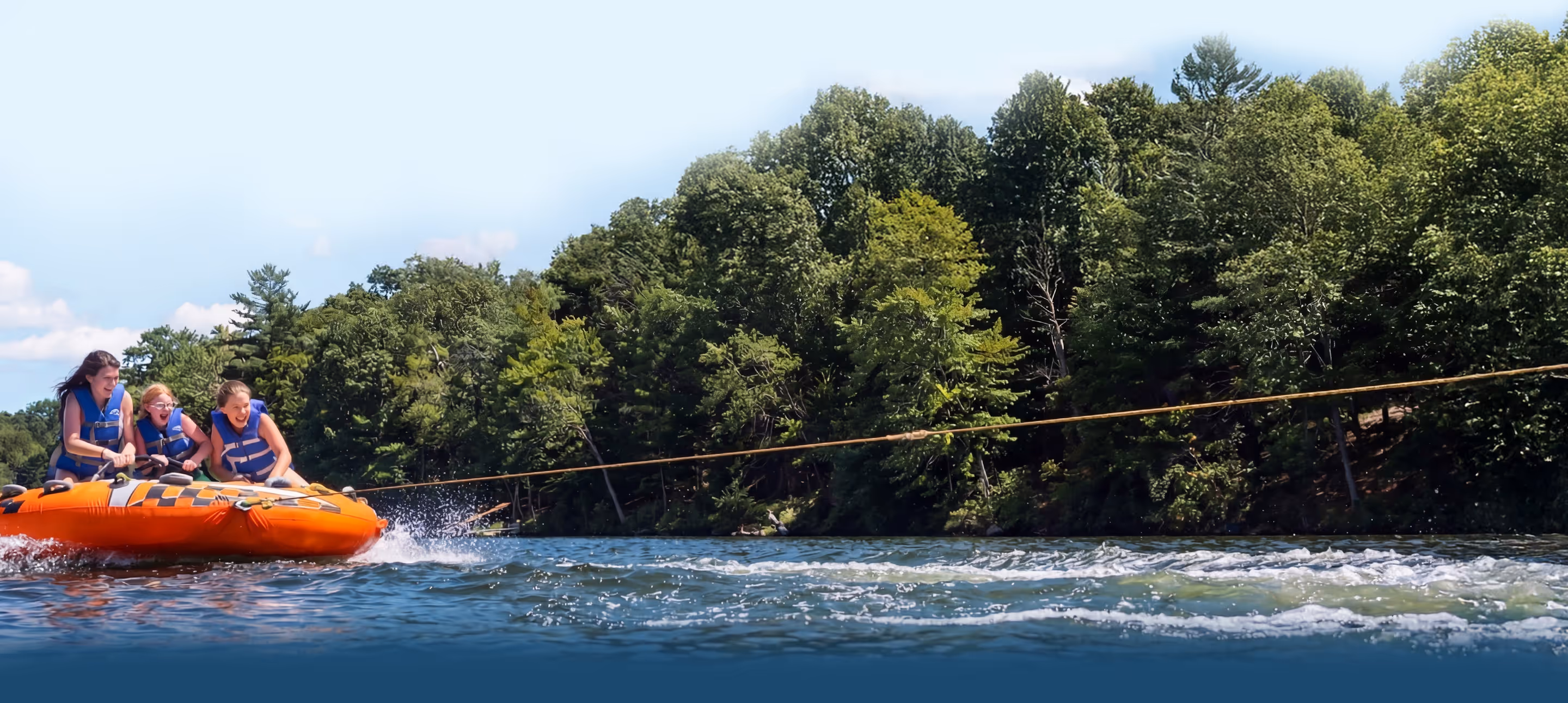 Three campers wearing life jackets riding an orange inflatable tube being pulled across Lake Hopatcong near a forested shoreline.