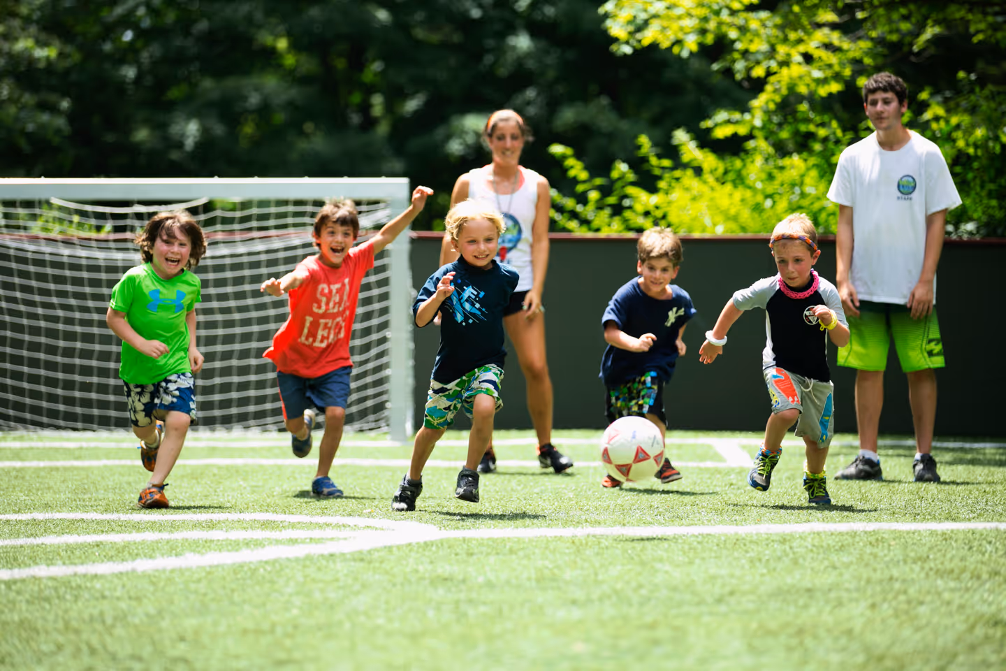 Group of young boys running and chasing a soccer ball on a green field with two counselors watching in the background.