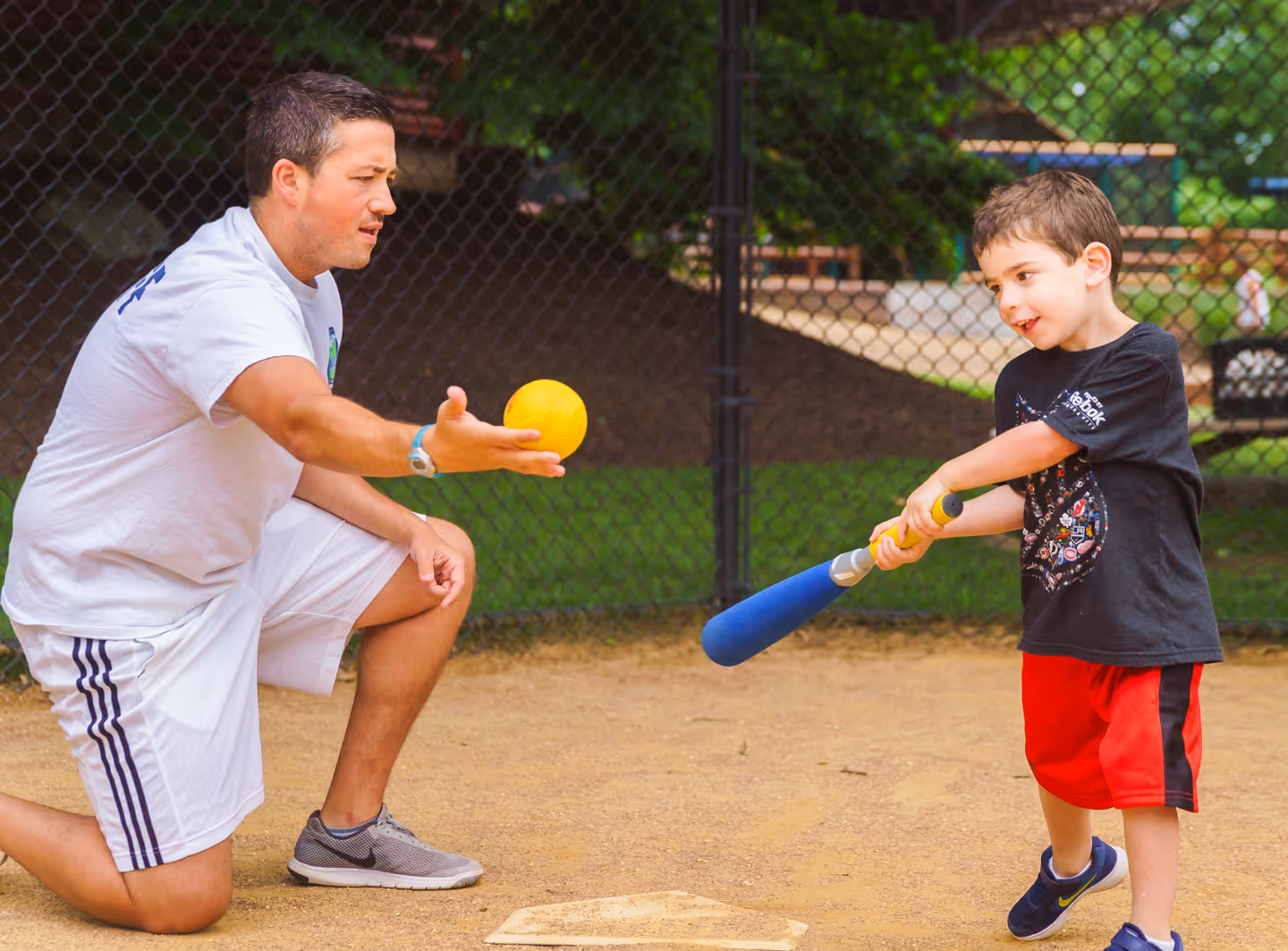 Young boy in red shorts holding a blue bat practicing hitting a yellow ball thrown by an adult man kneeling on a baseball field.