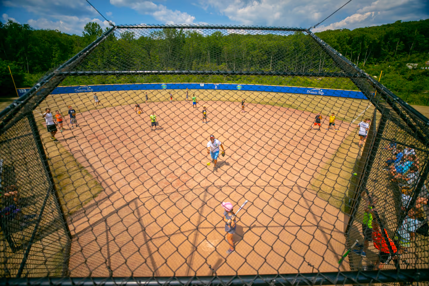 Overhead view of a youth softball game in progress on a dirt field surrounded by green trees and a blue outfield fence.