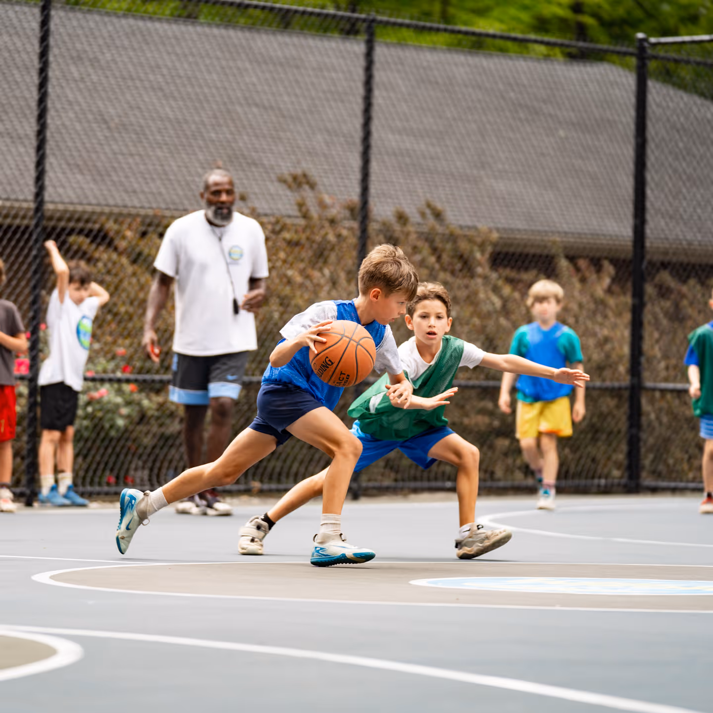 Two boys playing basketball on an outdoor court while a coach and other children watch in the background.