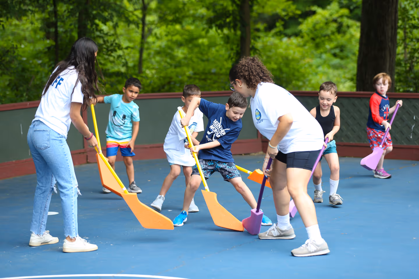 Children playing hockey with foam sticks on an outdoor court, supervised by two staff members.