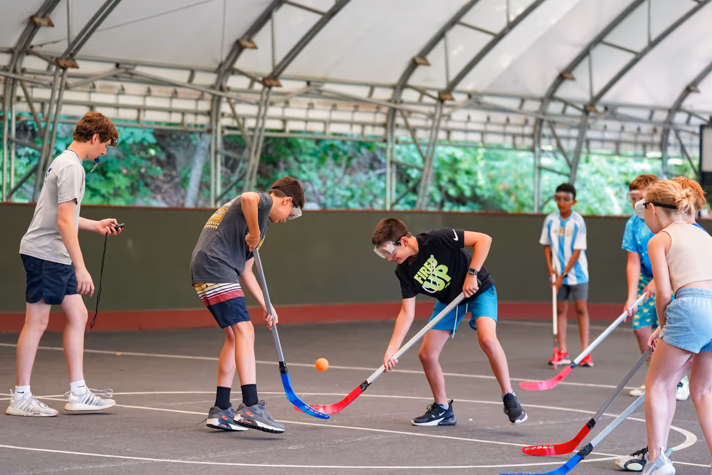 Group of kids playing floor hockey under a tented structure, two boys facing off at the center with a ball and stick.