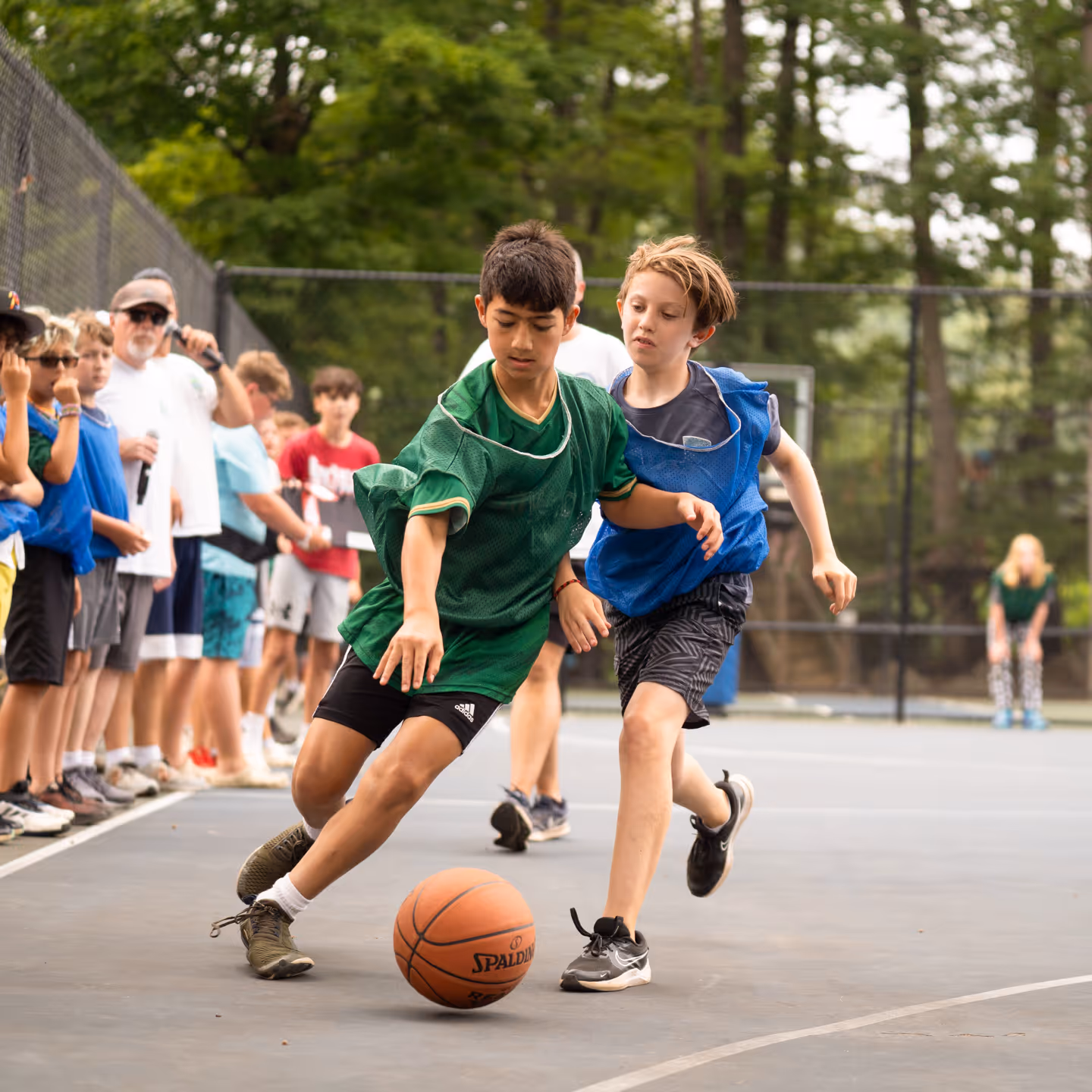 Two boys playing basketball on an outdoor court, one in a green jersey dribbling the ball while the other in a blue jersey defends.