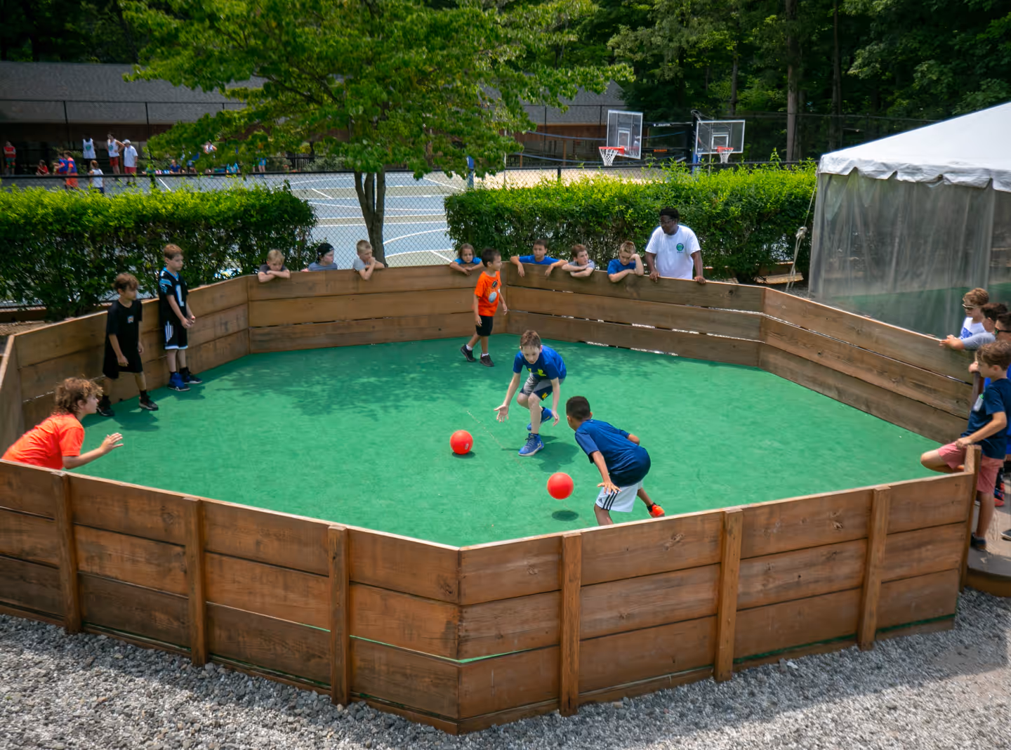 Children playing gaga ball inside a wooden pit with others watching around the edges.