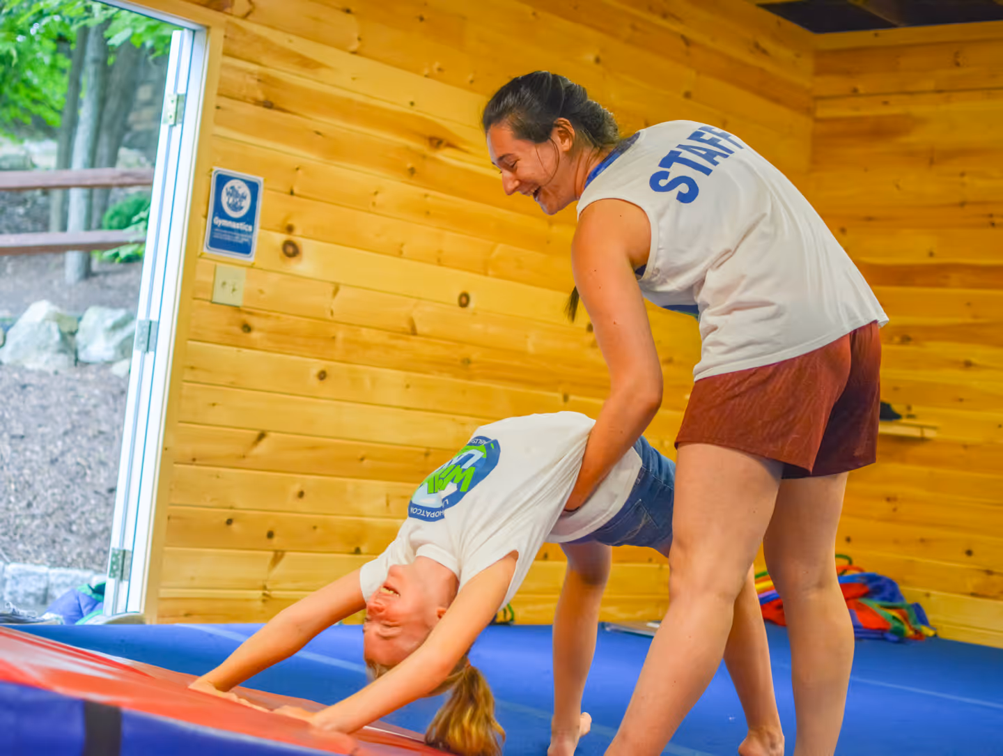 Female gymnastics staff member assisting a young girl performing a backbend inside a wooden gym.