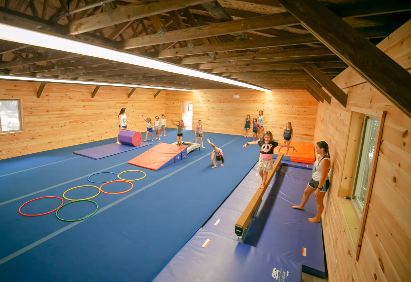 Indoor gymnastics facility with children practicing on balance beam, mats, and hoops under wooden beam ceiling.