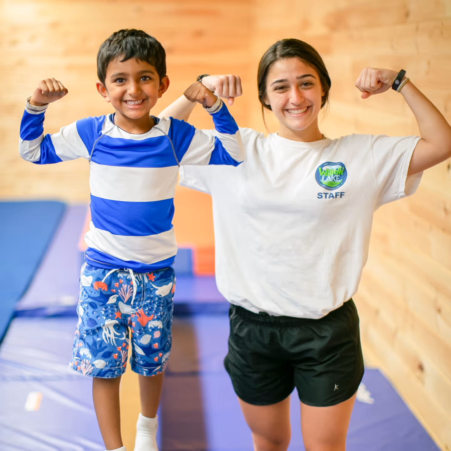 Smiling boy and female camp staff flexing their arm muscles indoors on blue gym mats.