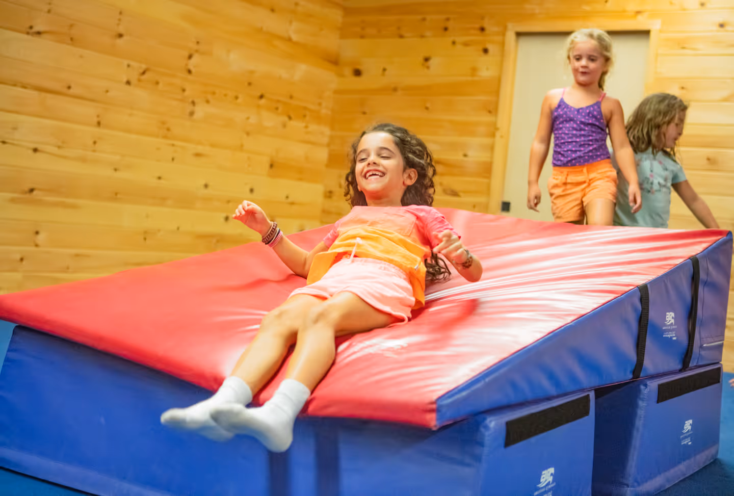 Young girl sliding down a red and blue gymnastics mat while two other children wait nearby.