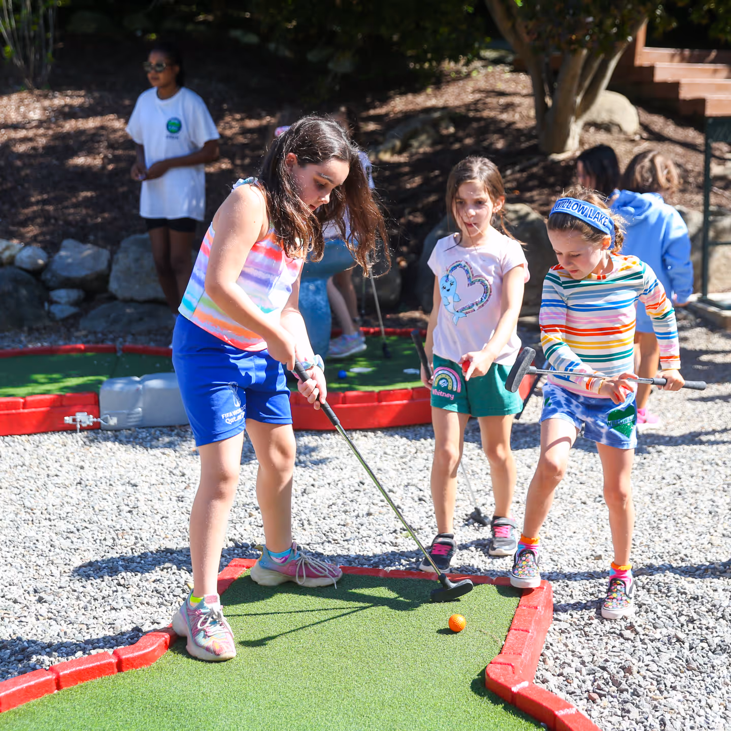 Three girls playing mini golf on a sunny day, with one girl preparing to putt an orange golf ball.