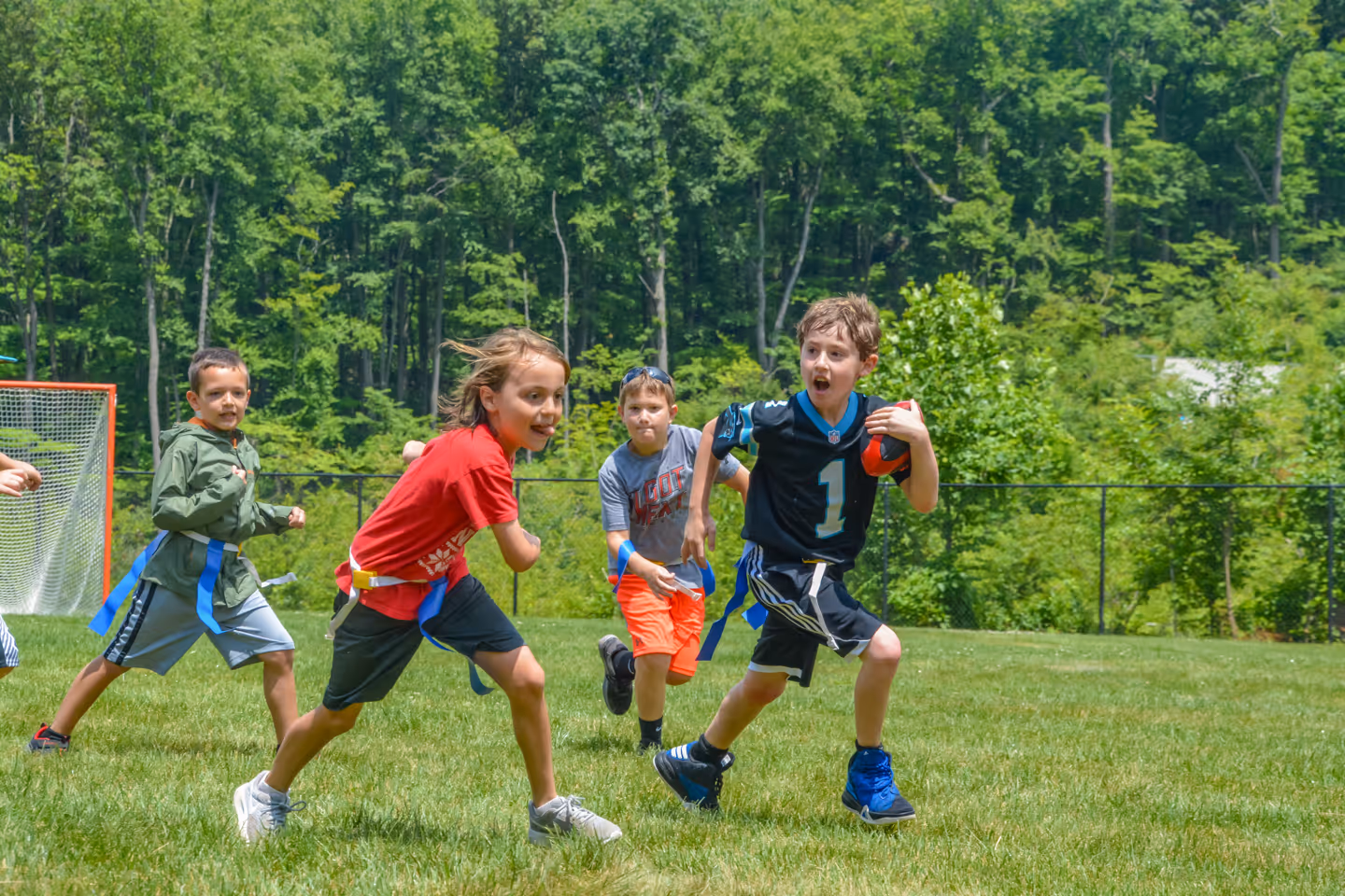 Group of children playing flag football outdoors on a sunny day with one boy running with the ball while others chase him.