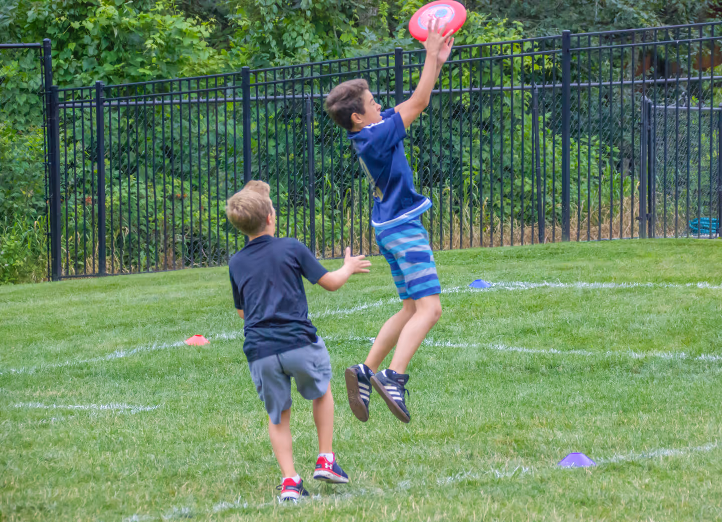 Two boys playing ultimate frisbee on grass with one boy jumping to catch a red frisbee.