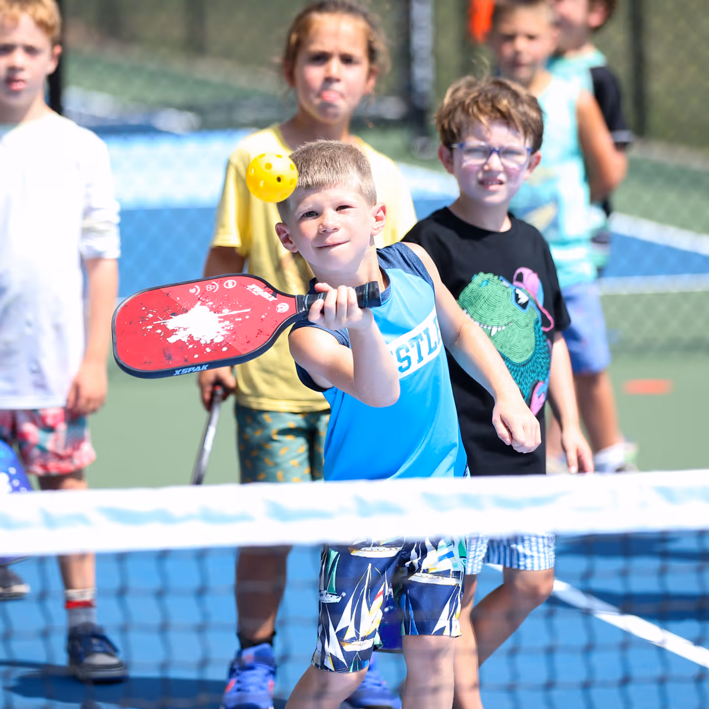 Young boy focused on hitting a yellow pickleball with a red paddle on a pickleball court, with other children watching behind him.