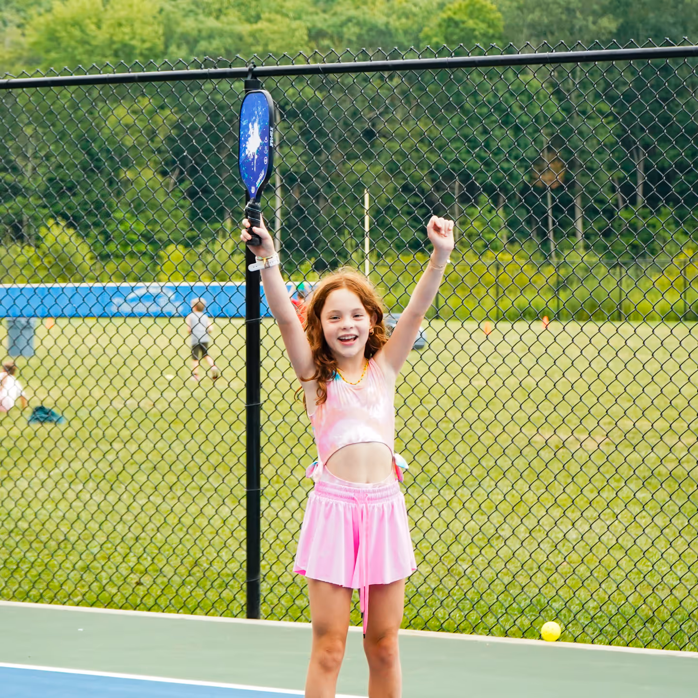 Young girl in pink sportswear celebrating with arms raised holding a pickleball paddle on an outdoor court.
