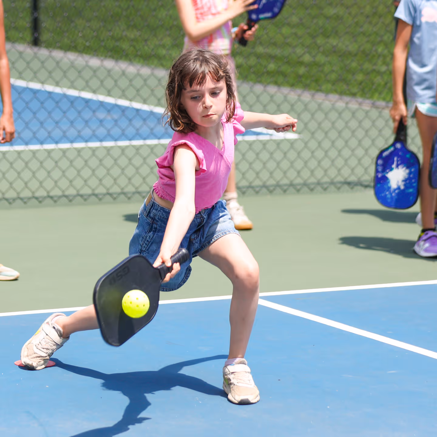 Young girl in a pink shirt and denim shorts reaching to hit a yellow pickleball with a black paddle on an outdoor court.