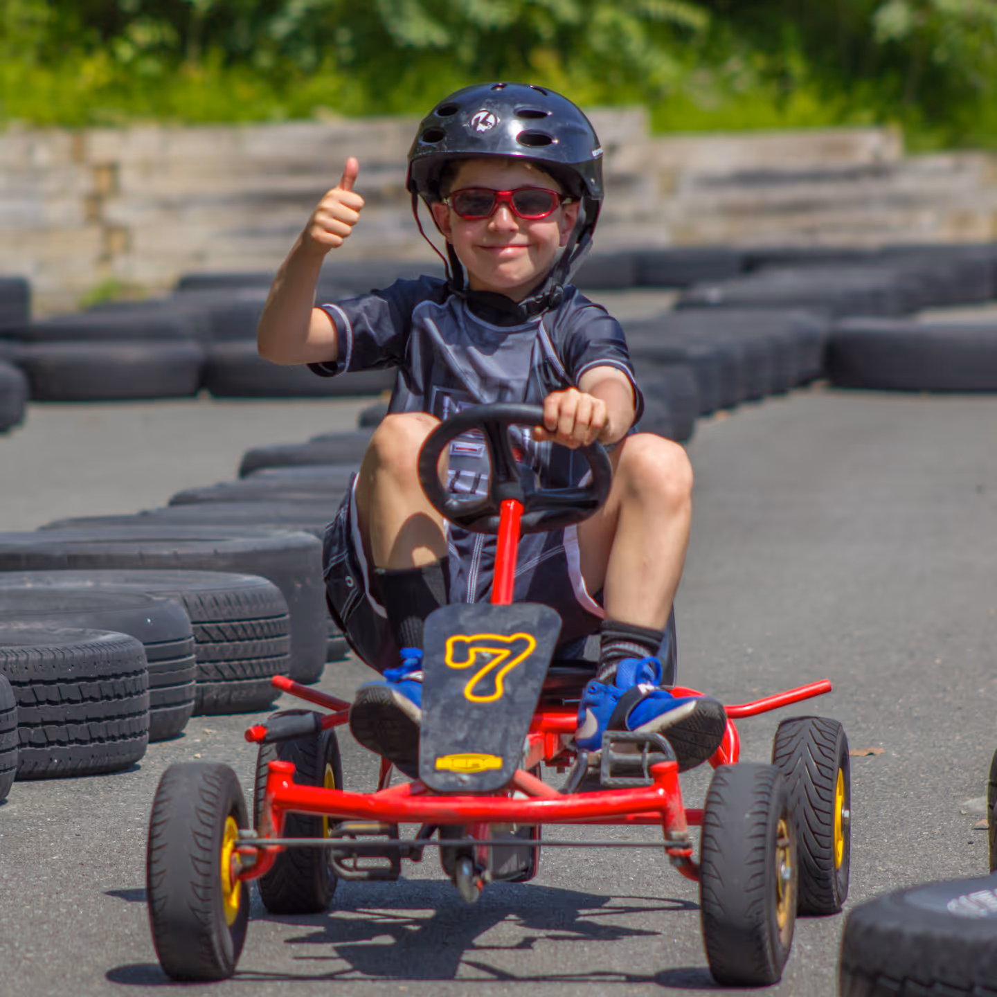 Boy wearing helmet and sunglasses giving thumbs up while riding a red pedal kart on a track lined with tires.