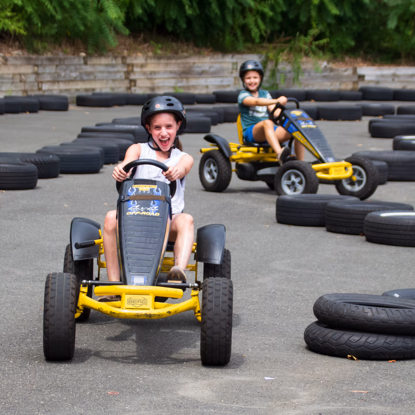 Two children wearing helmets riding yellow pedal go-karts on a paved track bordered by tires.