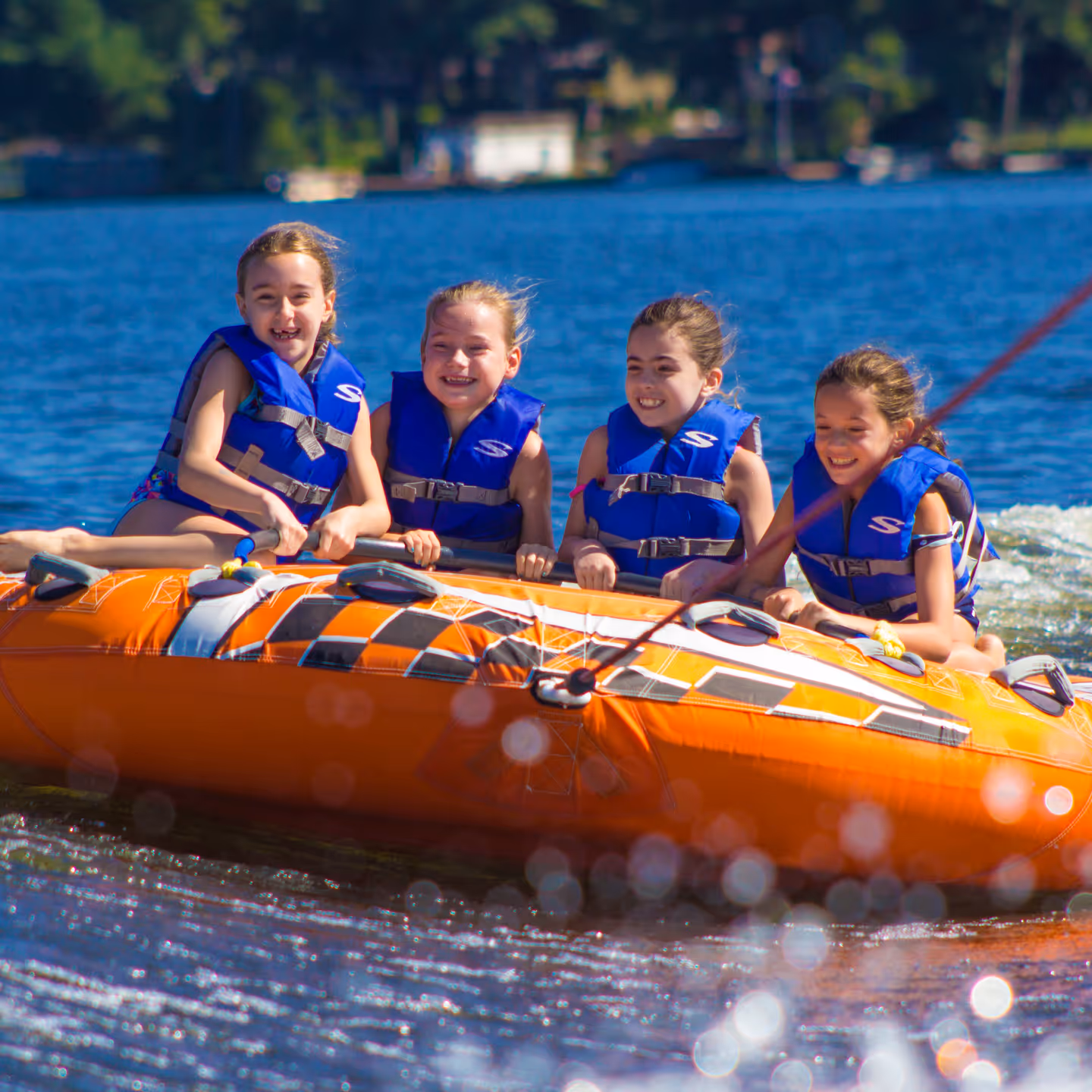 Four young girls wearing blue life jackets smiling while tubing on an orange inflatable tube on a lake.