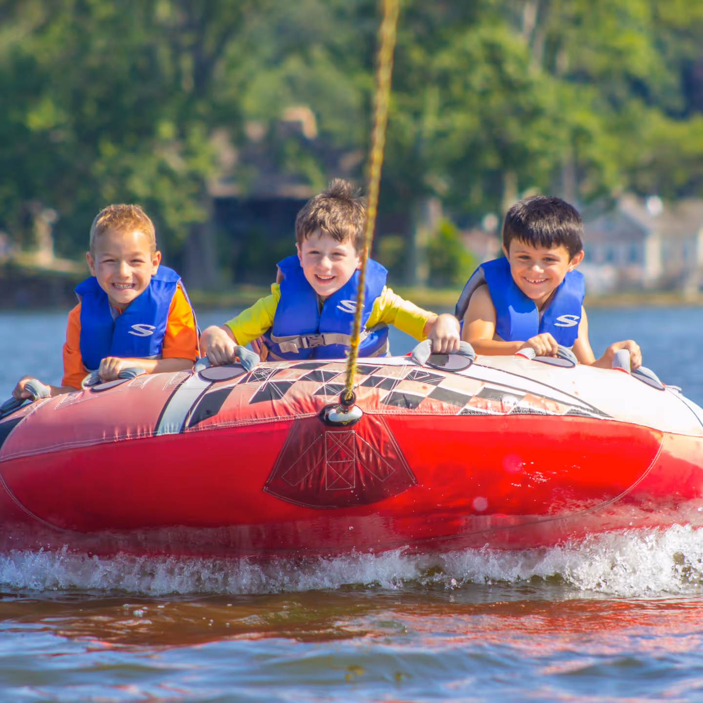 Three young boys wearing blue life jackets smiling while tubing on a lake.