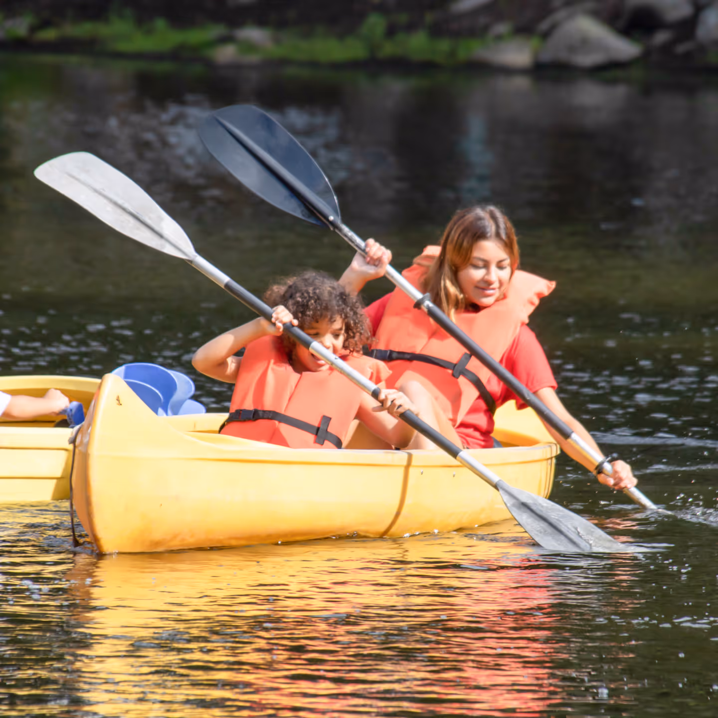 Two people wearing orange life jackets paddling a yellow canoe on calm water.