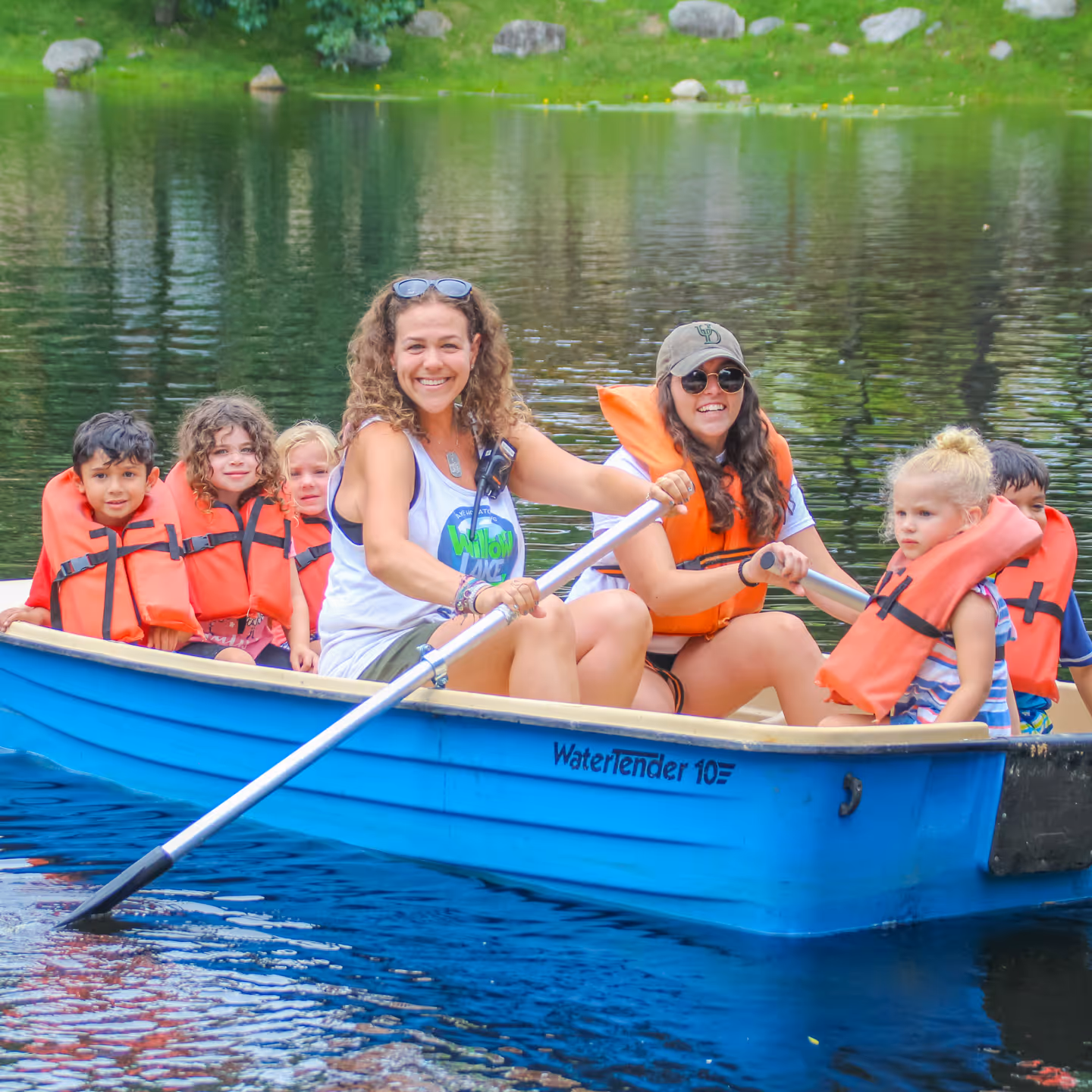 Two smiling women rowing a blue boat with six children wearing orange life jackets on a calm lake.