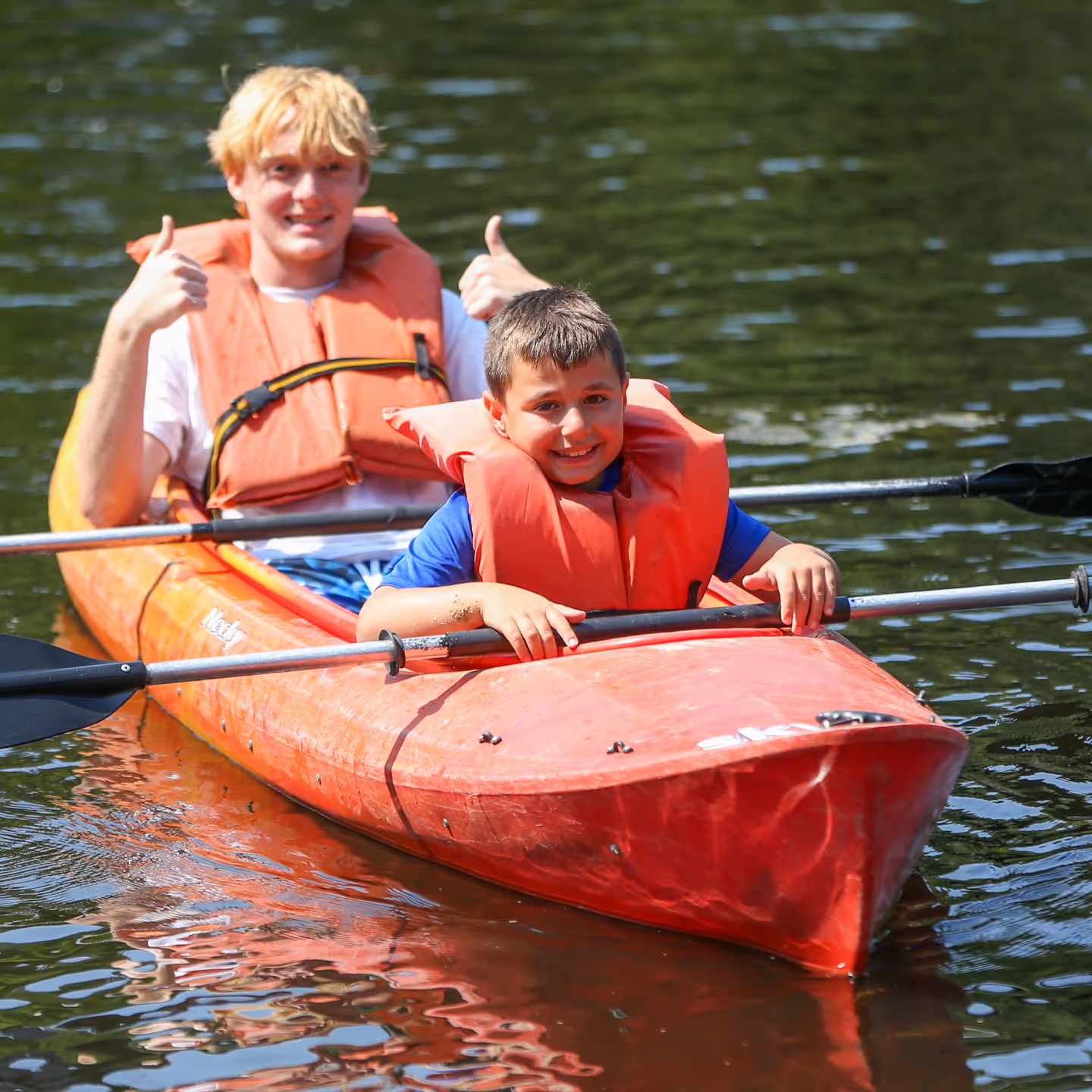 Two boys wearing orange life jackets smiling and sitting in a red kayak on calm water, one giving a thumbs up.