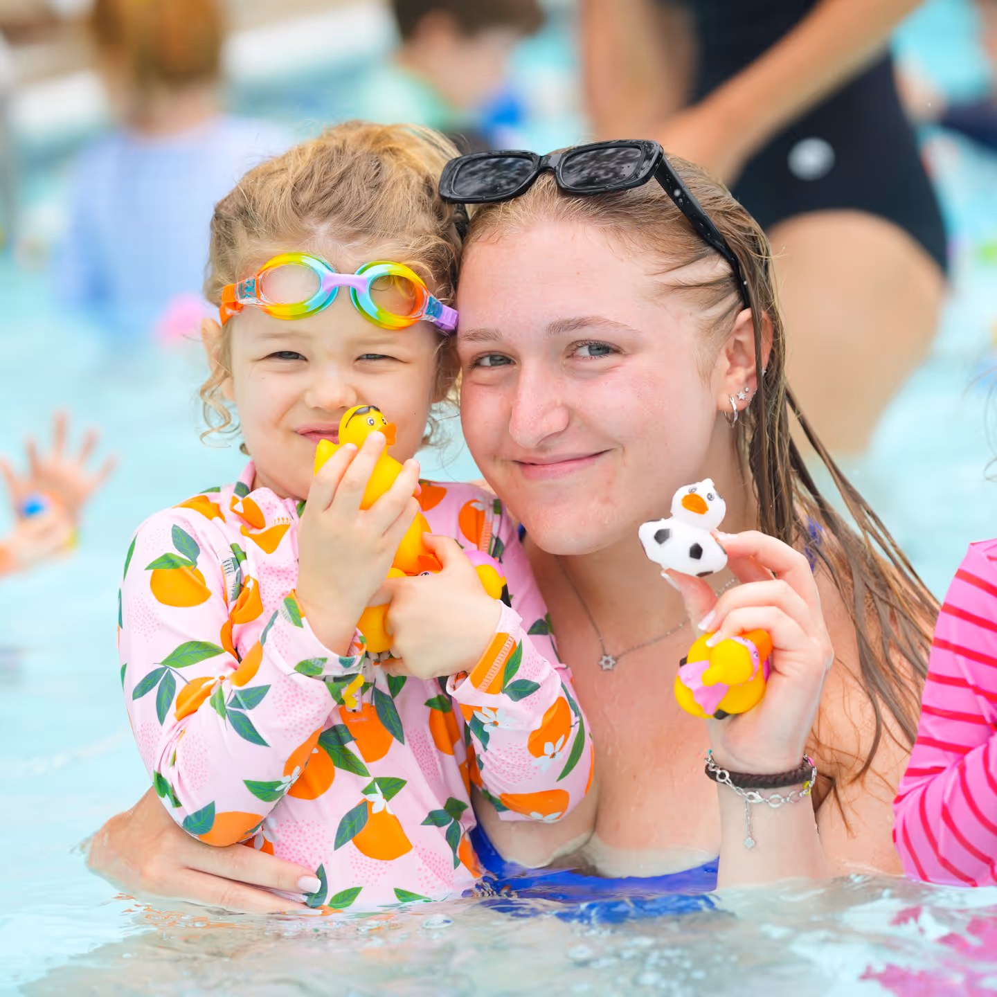 Smiling young woman and child in a pool holding rubber ducks, child wearing orange-themed swimwear and colorful goggles.